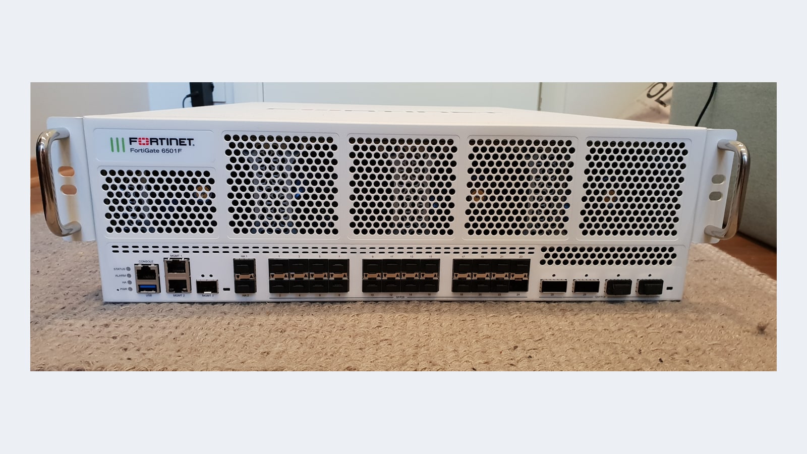 Vedere frontală a unui firewall Fortinet FortiGate 6501F alb, montabil în rack, pe un covor bej, cu grile de ventilație sus și rânduri de porturi SFP28 și QSFP28 jos; un dispozitiv FortiGate reprezentativ, nu unul dintre firewall-urile din intruziunea descrisă de Unit 42.