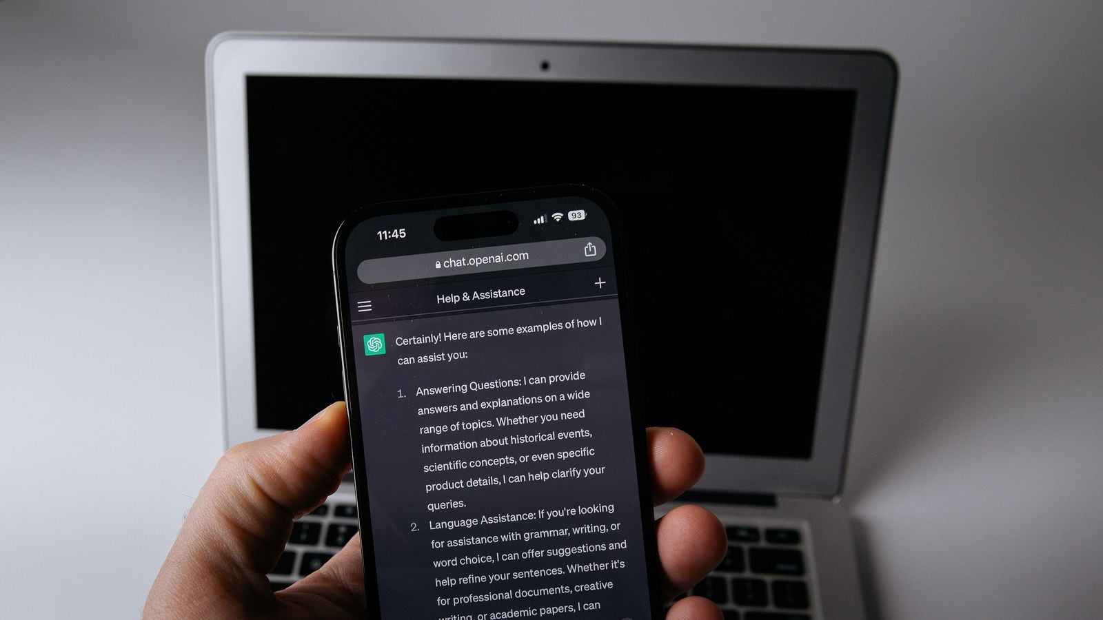 Une main tenant un smartphone qui affiche une conversation écrite avec ChatGPT dans un navigateur web mobile, devant un ordinateur portable ouvert